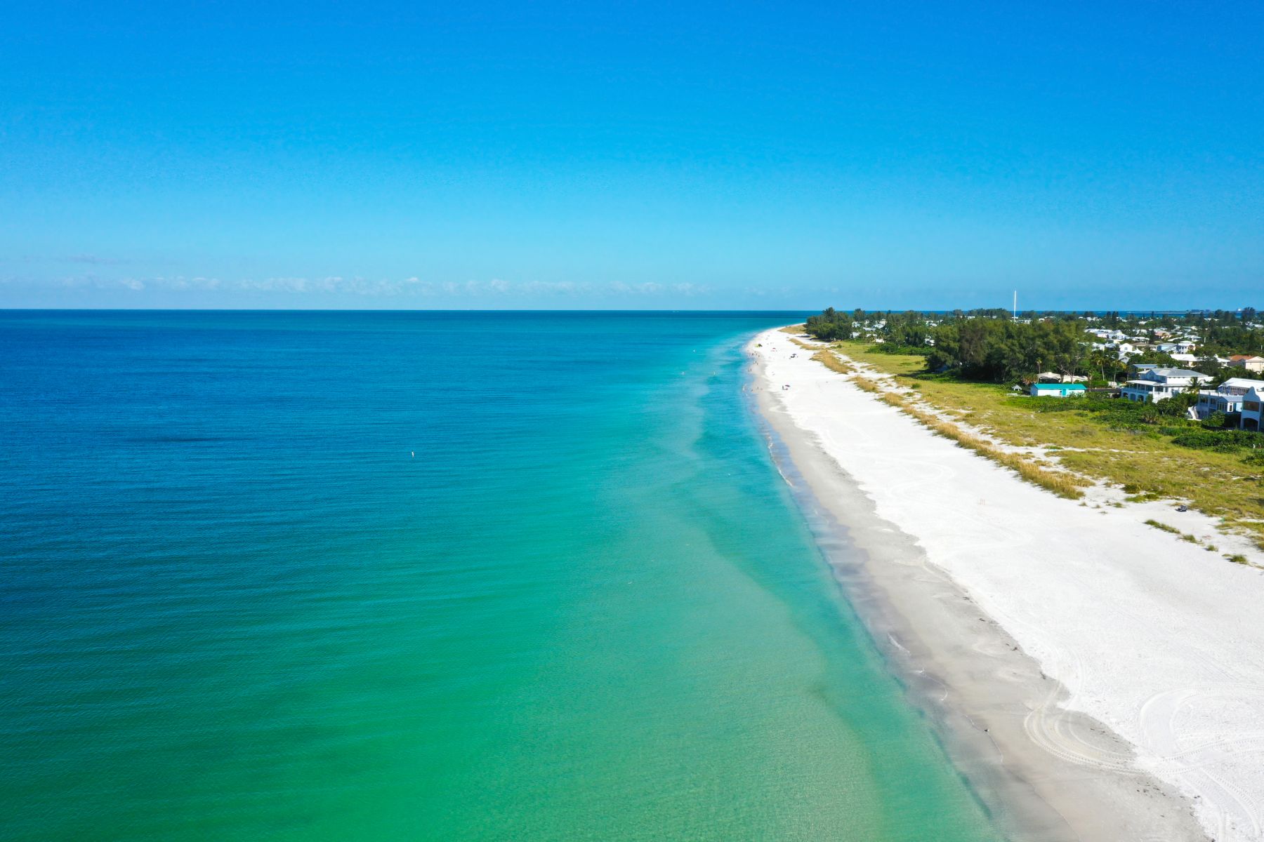 A stunning aerial shot showcasing the pristine white sand coastline and crystal-clear turquoise waters of Anna Maria Island under a bright blue sky. This panoramic view highlights the world-class beaches and tropical paradise atmosphere awaiting guests who book their stay with A Paradise Vacation Rentals.