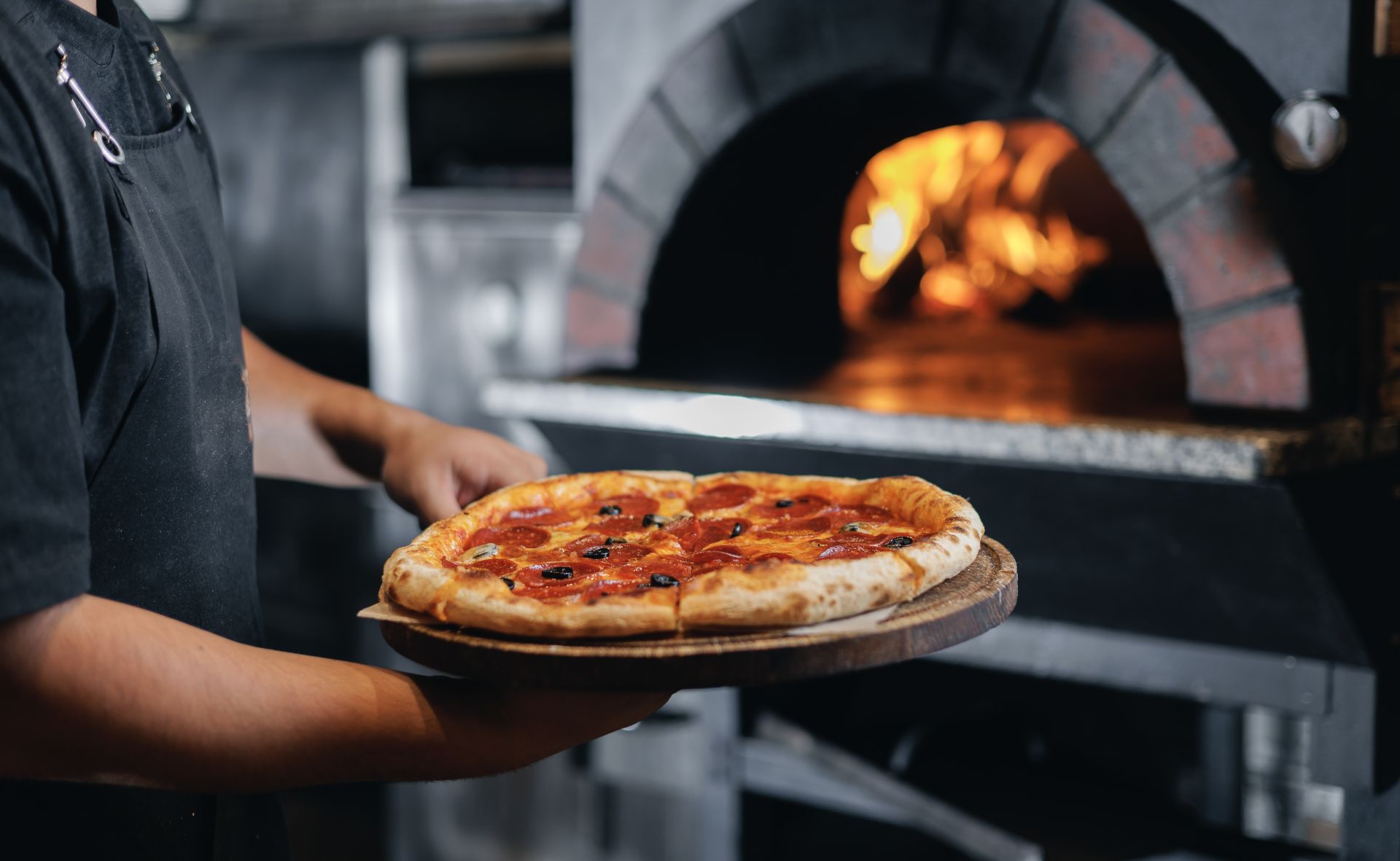 A chef holds a fresh, hot pepperoni and olive pizza on a wooden peel in front of a glowing wood-fired brick oven. This appetizing scene highlights the gourmet dining experiences and local flavors available to guests staying with A Paradise Vacation Rentals on Anna Maria Island.